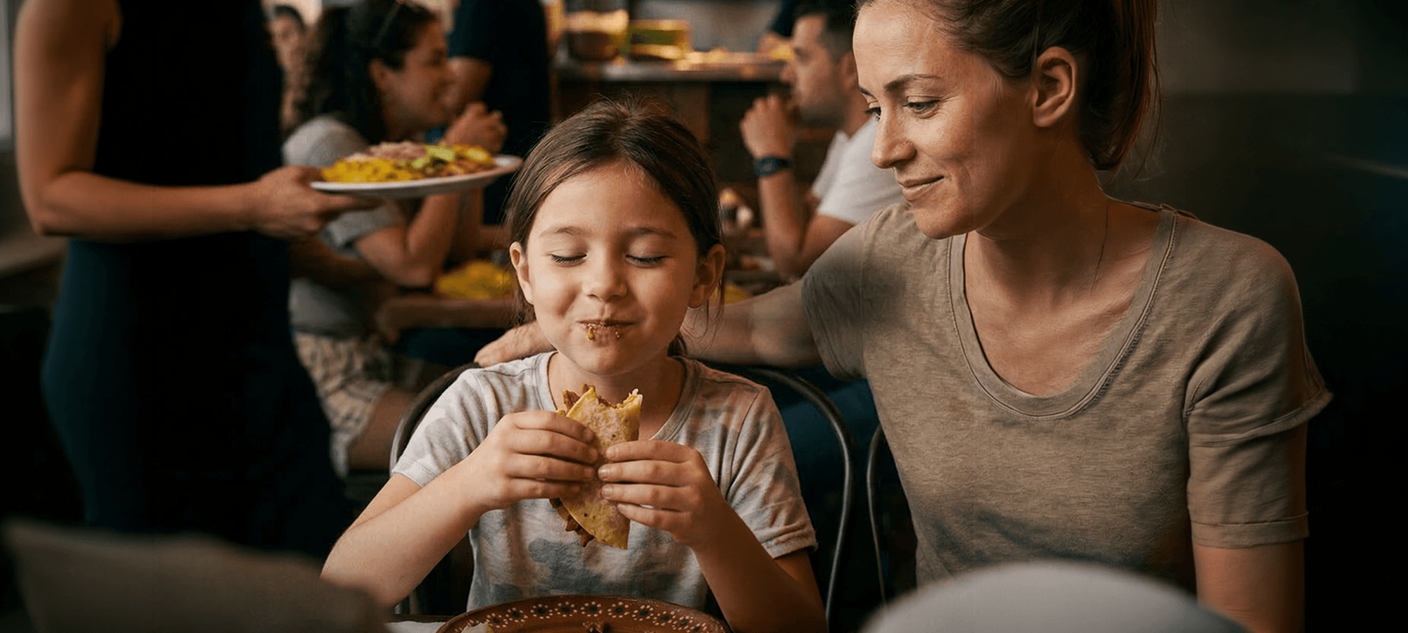 Girl enjoys eat taco safely while mother watches smiling